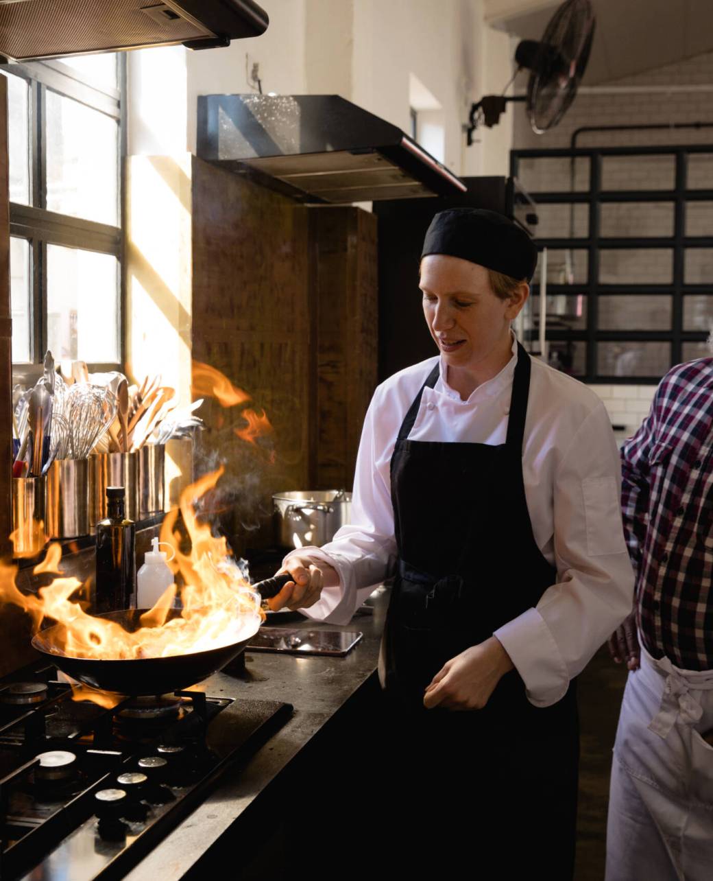 Side view of a Caucasian woman and an African man at a cookery class, frying vegetables on a frying pan, flaming