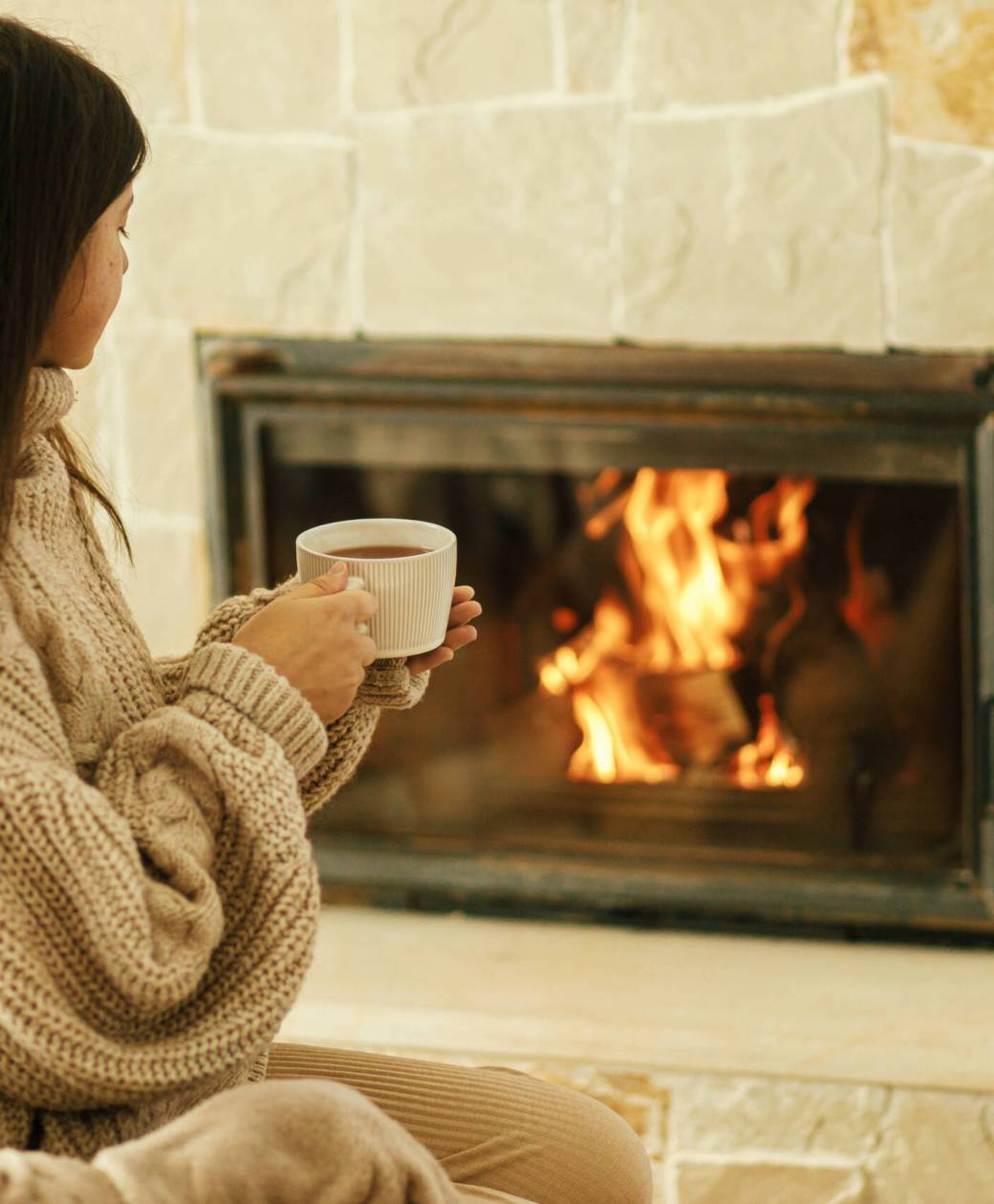 Woman in cozy sweater holding cup of warm tea at fireplace in rustic room. Heating house in winter with wood burning stove. Young stylish female relaxing at fireplace in farmhouse