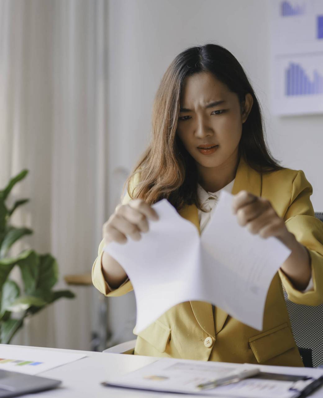 Young businesswoman is experiencing extreme frustration while working in her office and tearing a document in half